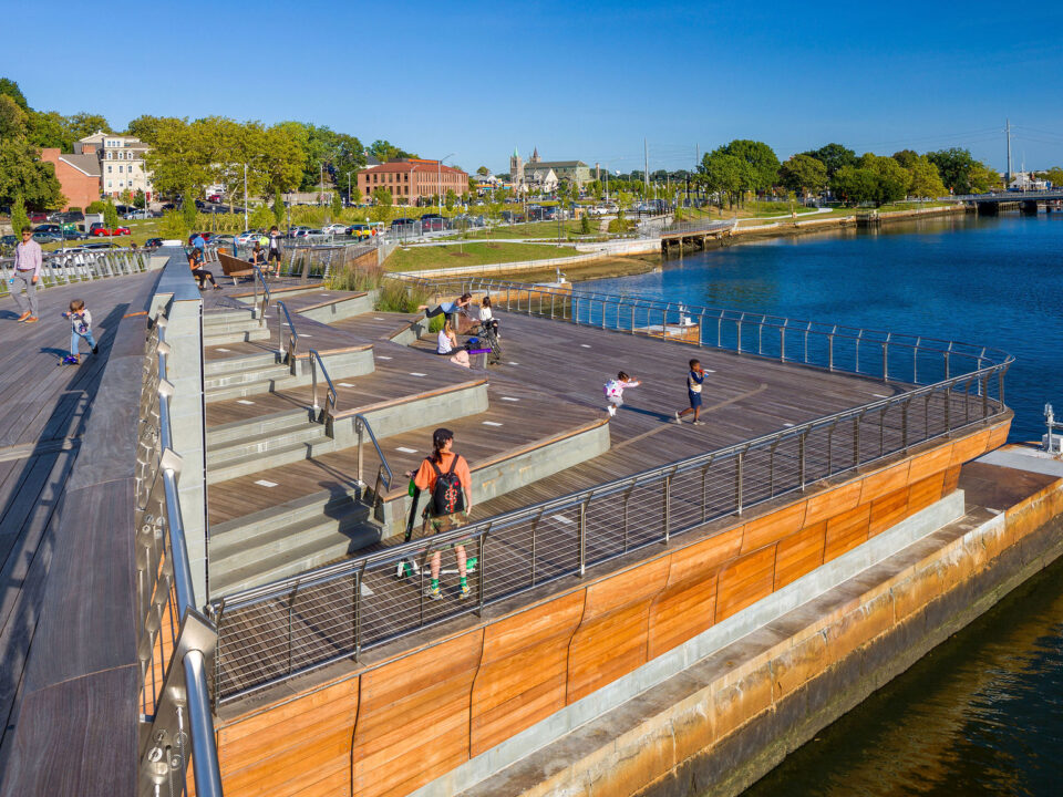 People are walking along the Michael S. Van Leesten Memorial Bridge near a river.