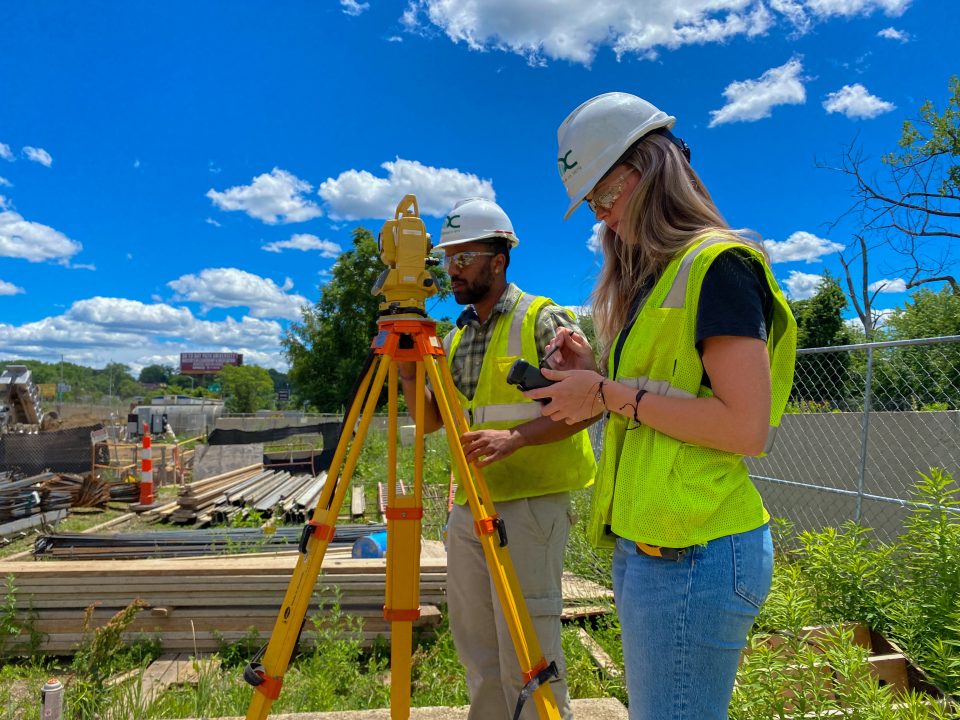 Two students pursuing careers in construction collaborate at a site, utilizing a surveyor's tool.
