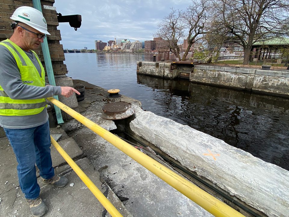 A man, committed to safety, is standing next to a body of water wearing a yellow vest.